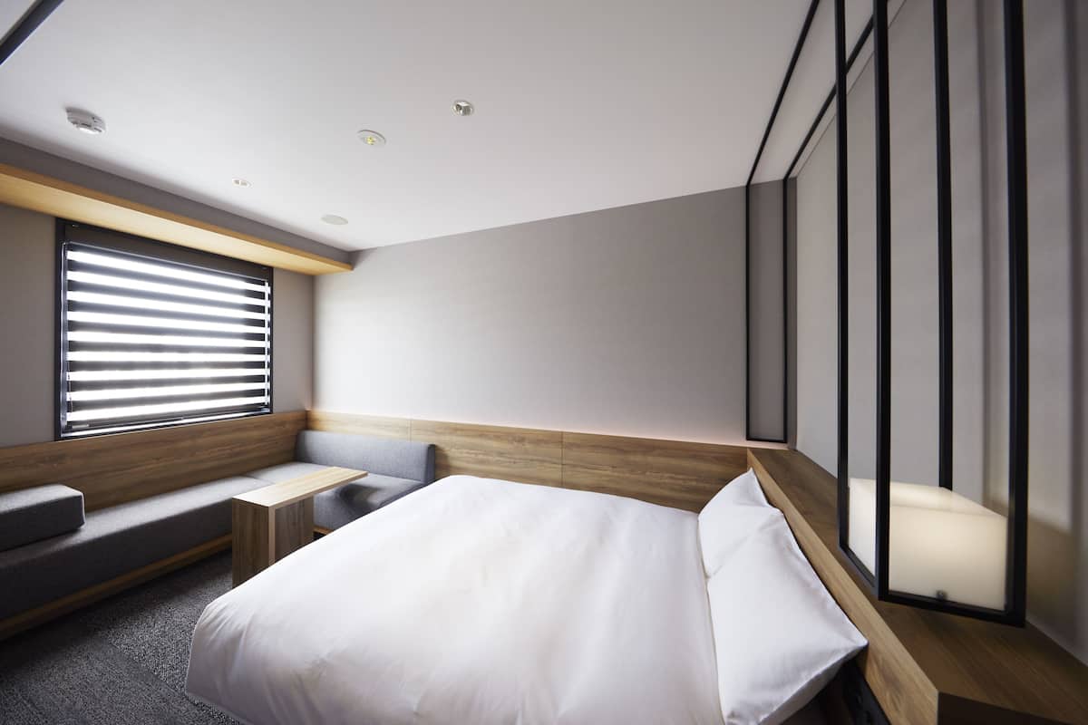 A modern hotel room with a large bed, built-in bench seating, a small table, and a large window with blinds.