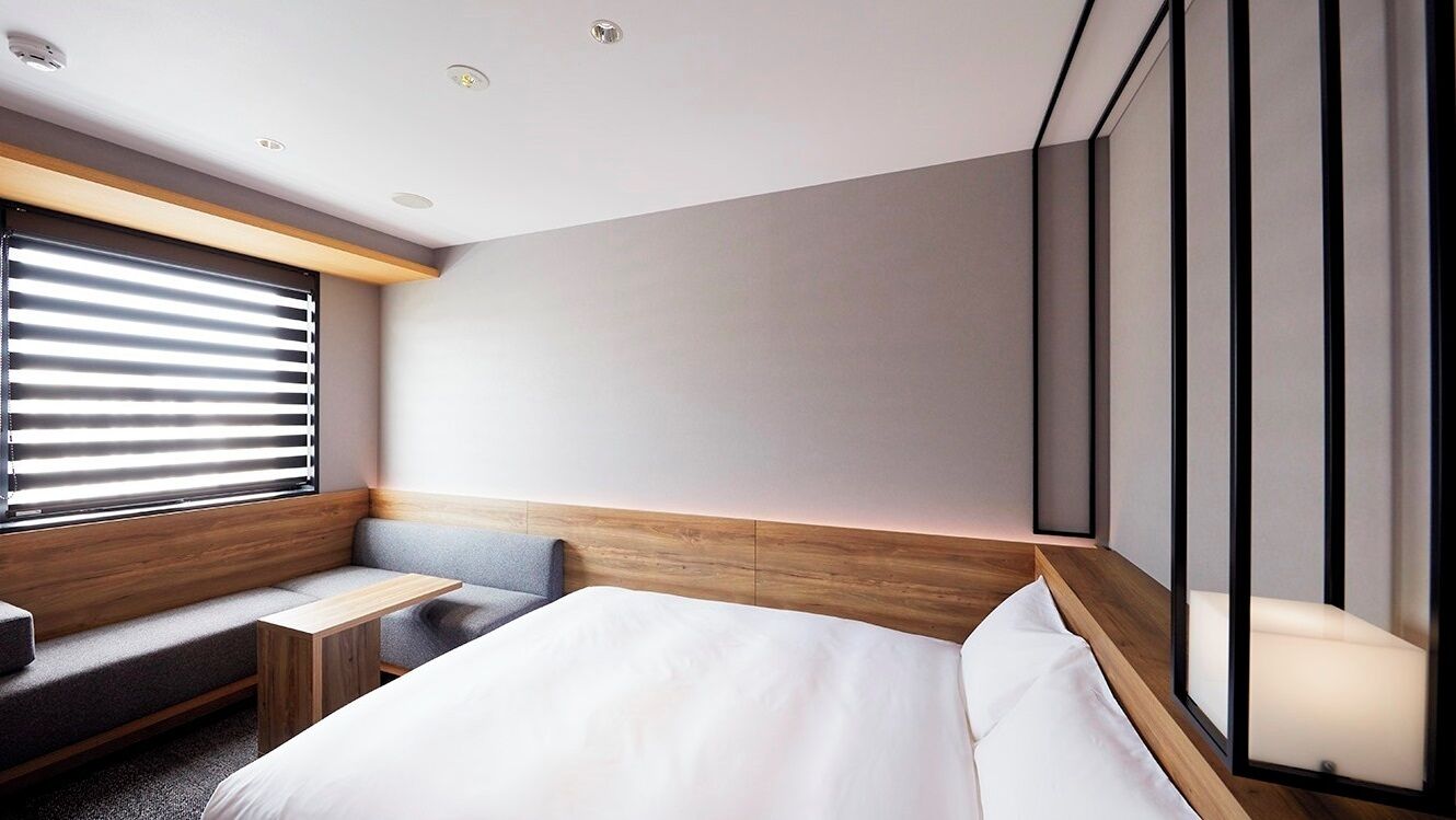 A modern hotel room with a bed, a seating area, a small table, and a large window with blinds.