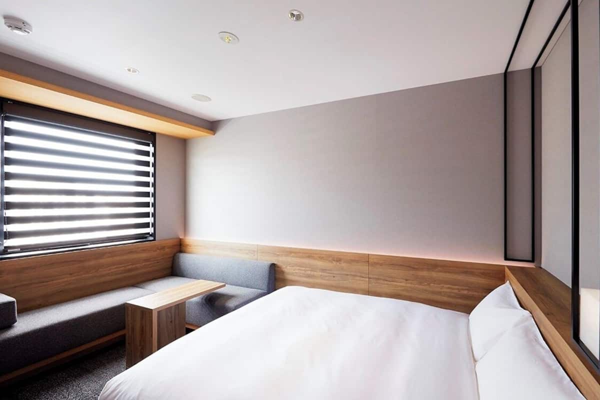 A modern hotel room with a bed, a seating area, a small table, and a large window with blinds.