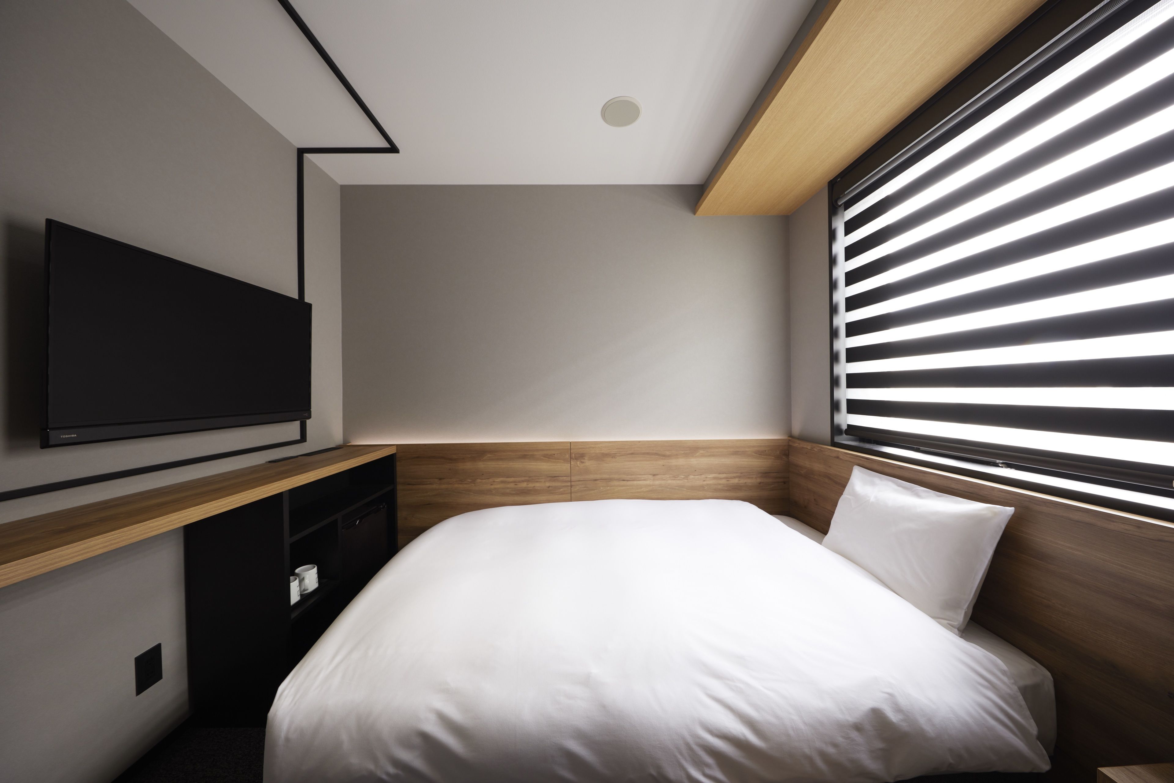 A modern hotel room with a large bed, a flat-screen TV mounted on the wall, and a window with horizontal blinds.