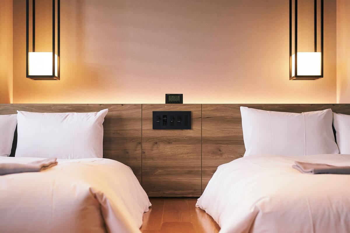 A hotel room with two beds, a wooden headboard, a digital clock, and wall-mounted lights.