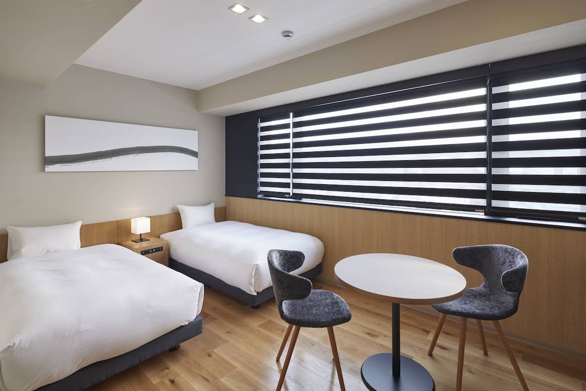 Une chambre d’hôtel avec deux lits, une table ronde avec des chaises, une fenêtre avec des stores et un tableau au mur.