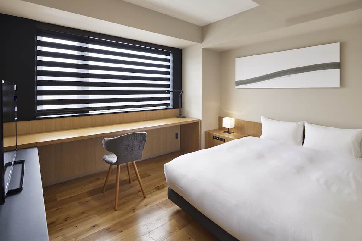 Une chambre d’hôtel moderne dotée d’un grand lit, d’un bureau avec une chaise, d’une table de chevet avec une lampe et d’une grande fenêtre avec des stores.