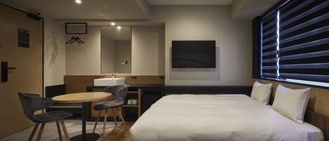 A hotel room with a bed, two chairs, a desk, a mirror, a TV, and a window with blinds.