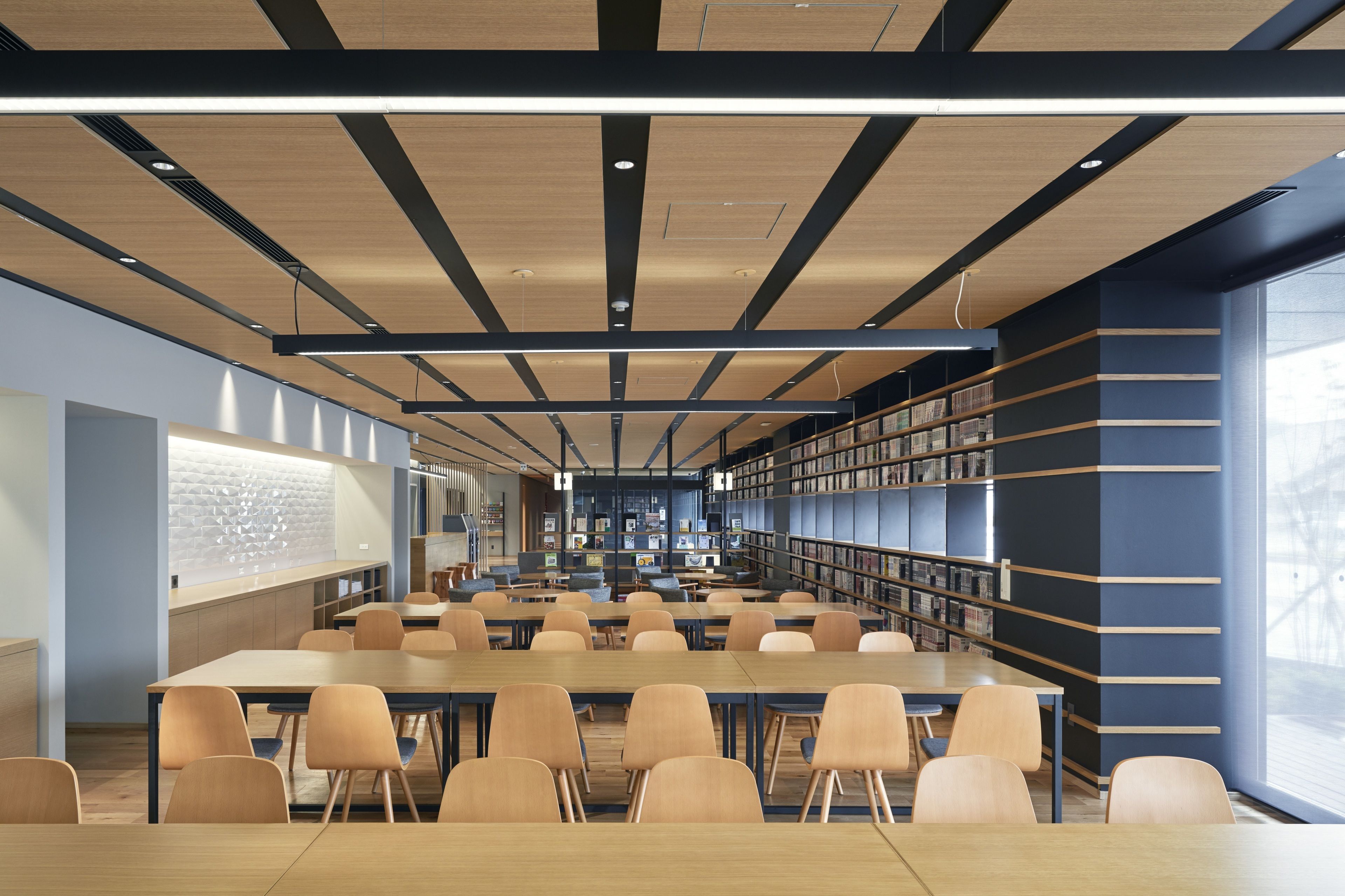 A modern library with long tables and chairs, bookshelves, and a large window.