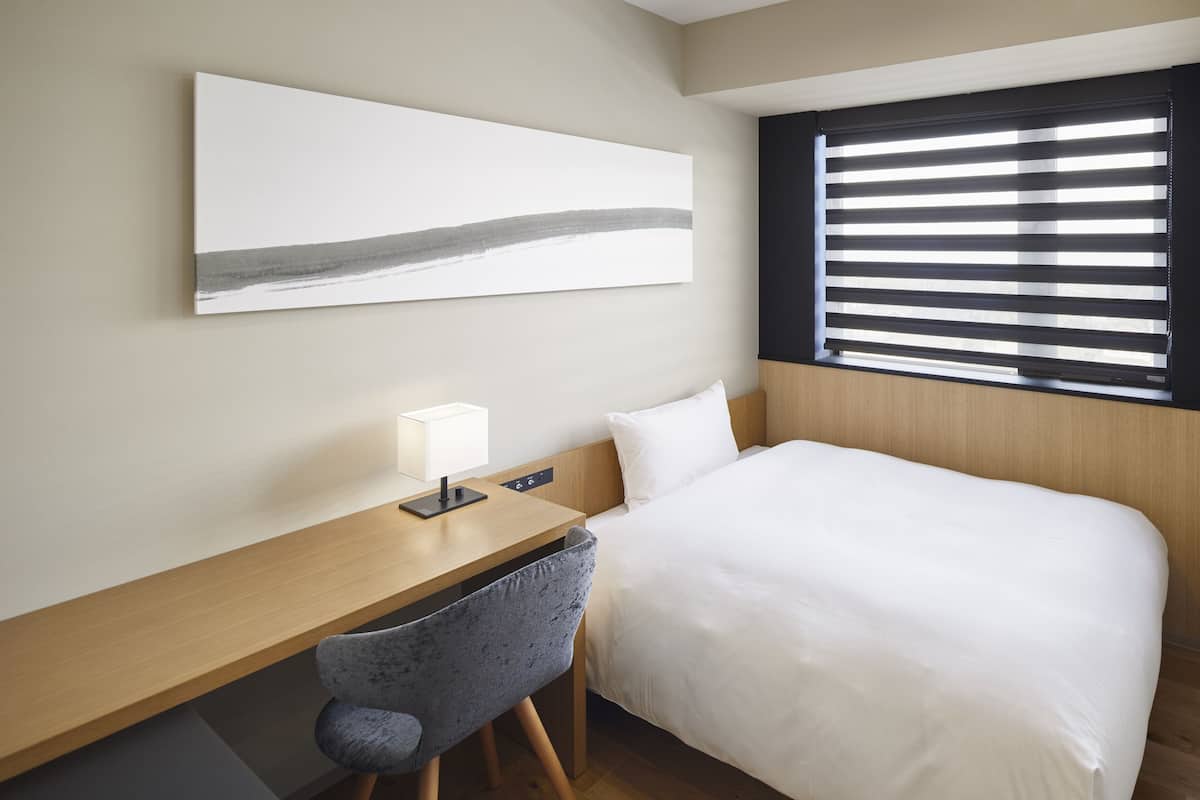 Une chambre d’hôtel comprenant un lit, un bureau, une chaise, une table de chevet, une lampe, une fenêtre avec stores et une œuvre d’art accrochée au mur.