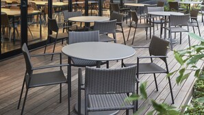 Un espace extérieur aménagé avec des tables et des chaises, la façade d’un bâtiment dotée de grandes fenêtres et une enseigne sur laquelle on peut lire « OBATAN ».
