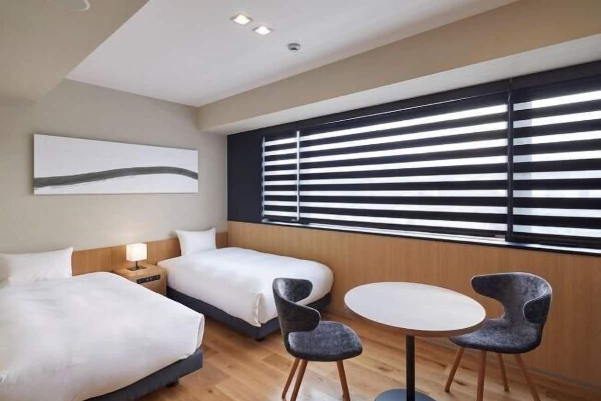 Une chambre d’hôtel moderne avec deux lits, une table à manger et une grande fenêtre avec des stores.