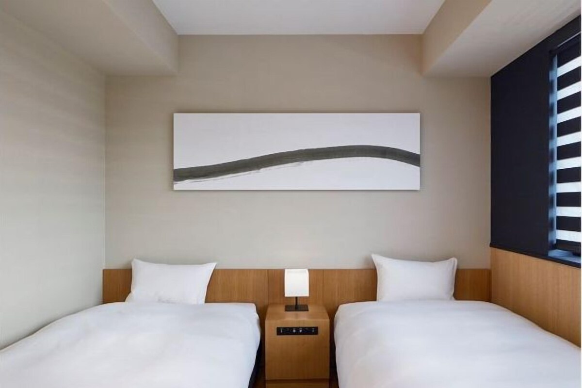 Habitación de hotel con dos camas, una mesita de noche con lámpara, un cuadro en la pared y una ventana con persianas.