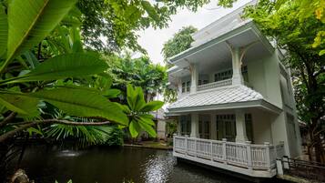 A two-story house with a balcony, surrounded by lush greenery and a water body.
