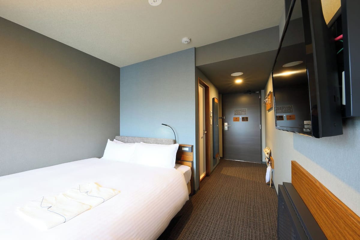 Een hotelkamer met een bed, een nachtkastje, een lamp, een spiegel en een deur die naar een gang leidt.