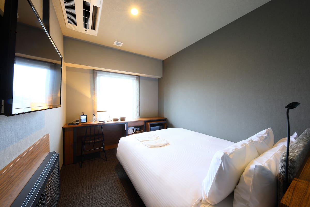 Een hotelkamer met een groot bed, een bureau met een computer, een stoel en een raam met jaloezieën.