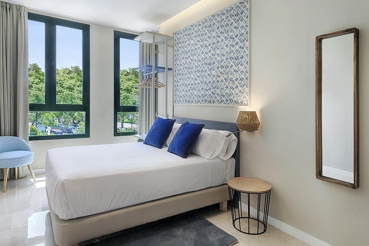 A modern bedroom with a large bed, two blue pillows, a bedside table with a lamp, a round side table, and a view of greenery through large windows.
