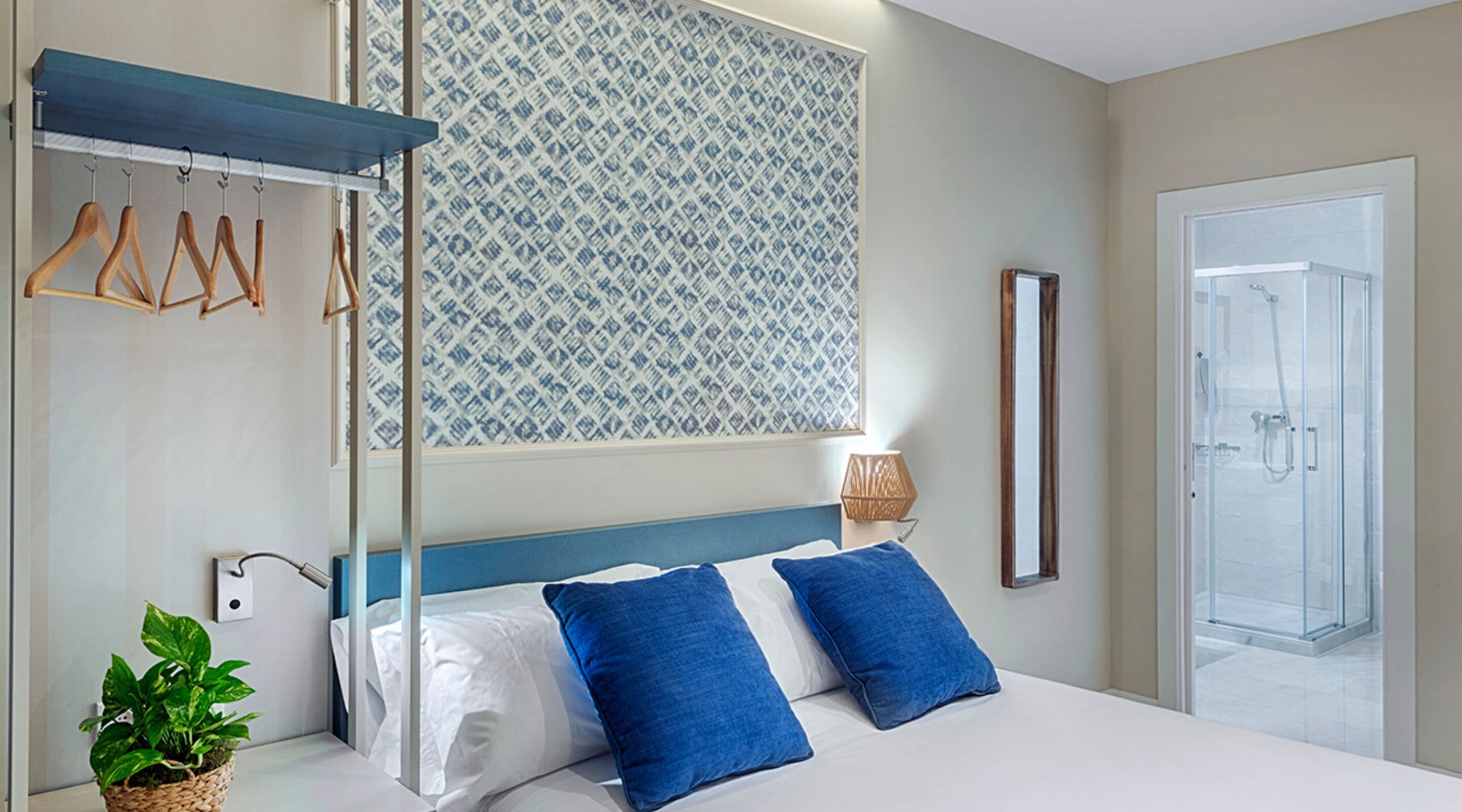 Una habitación de hotel moderna con una cama grande, un perchero, un ventanal con diseño, una lámpara de noche y un box de ducha de vidrio.