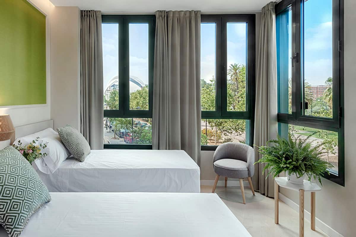 A hotel room with two beds, a chair, a small table with a plant, and large windows with curtains.