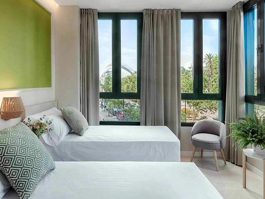 Habitación de hotel con dos camas, una silla, una mesita con una planta y ventanas amplias con cortinas.