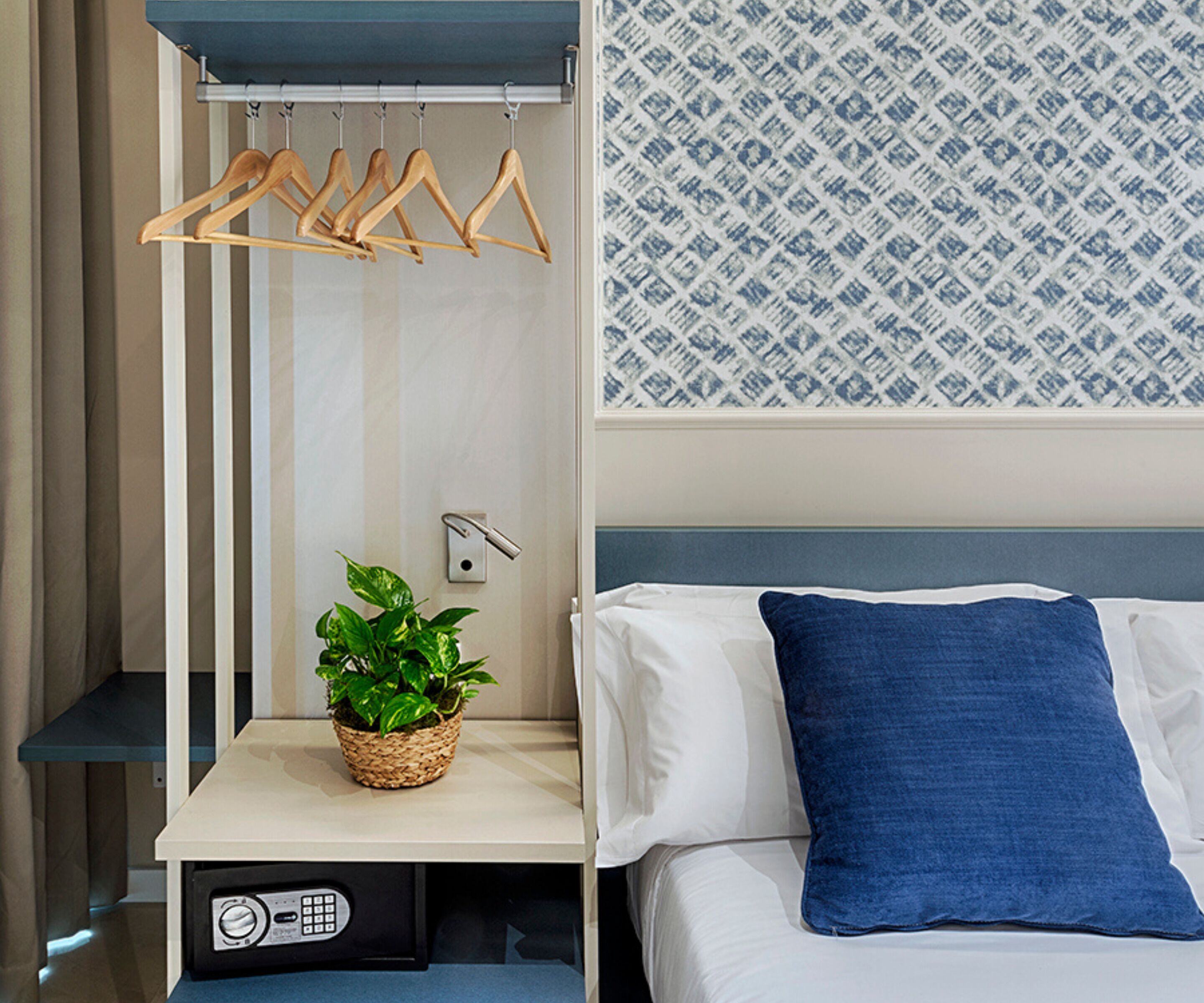 A hotel room with a bed, a bedside table with a plant, a hanging clothes rack, and a patterned wall.