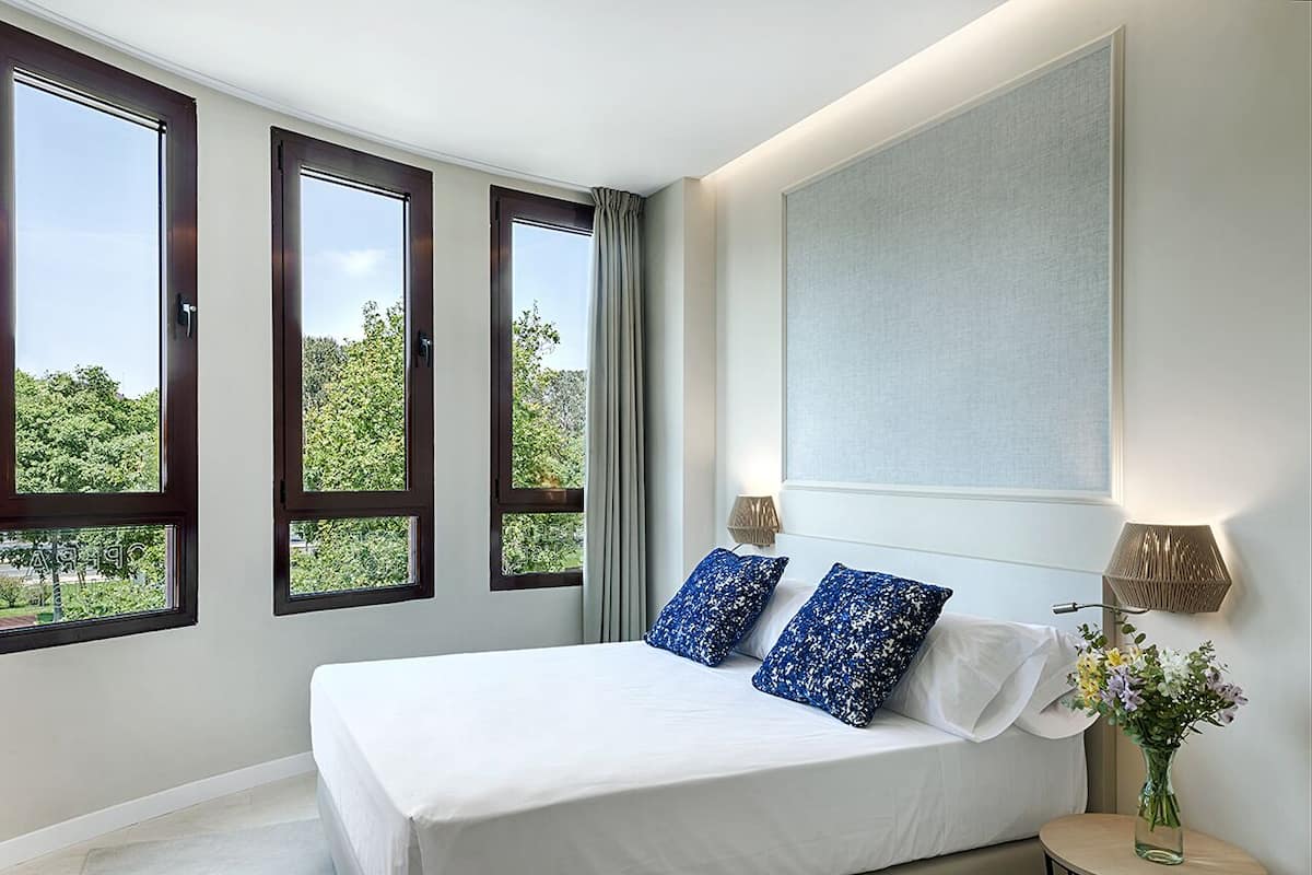 A bedroom with a large bed, two windows, a nightstand, and a wall-mounted lamp.