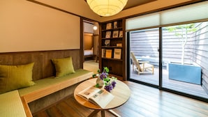 A room with a wooden bench, a small round table with a book and flowers, a bookshelf, and a sliding glass door leading to an outdoor area. - Saik (Kanazawa)