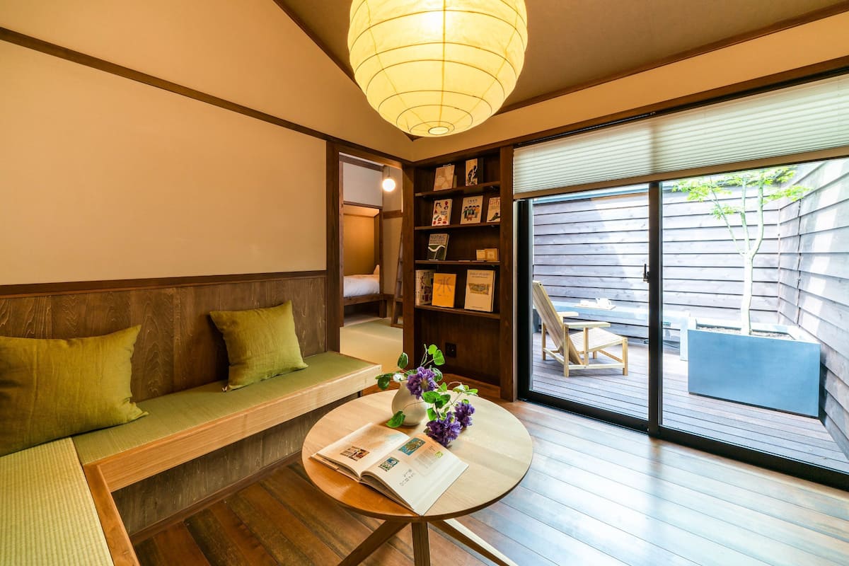 A room with a wooden bench, a small round table with a book and flowers, a bookshelf, and a sliding glass door leading to an outdoor area.