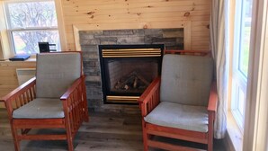 A cozy room with a stone fireplace, two wooden chairs, and a window with a view of trees.