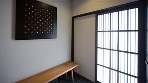 A hallway with a wooden bench, a wall-mounted light panel, and a sliding door with vertical blinds. - Higashiyama Aritsuki (Kanazawa)
