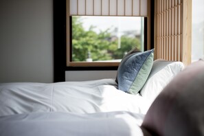 A neatly made bed with white linens and a pillow, a window with a view of greenery outside, and a wooden slat wall.