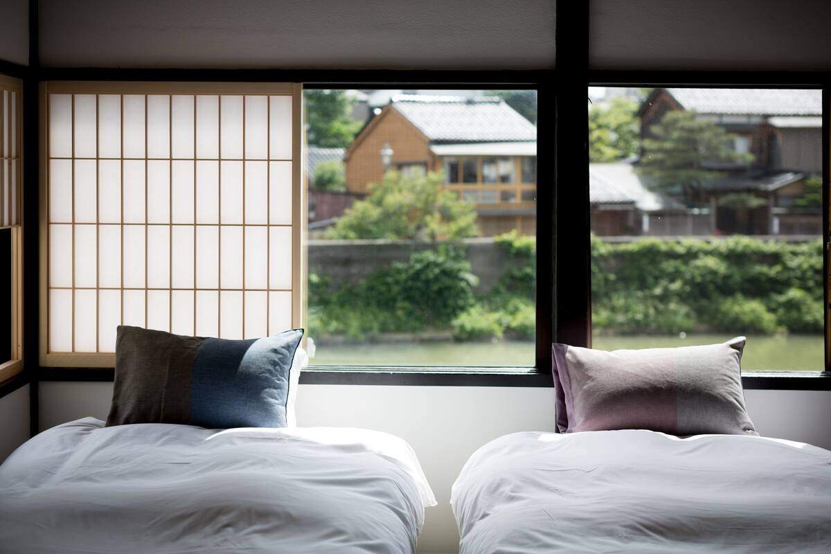 Quarto com duas camas, janela com vista para casas tradicionais e vegetação.