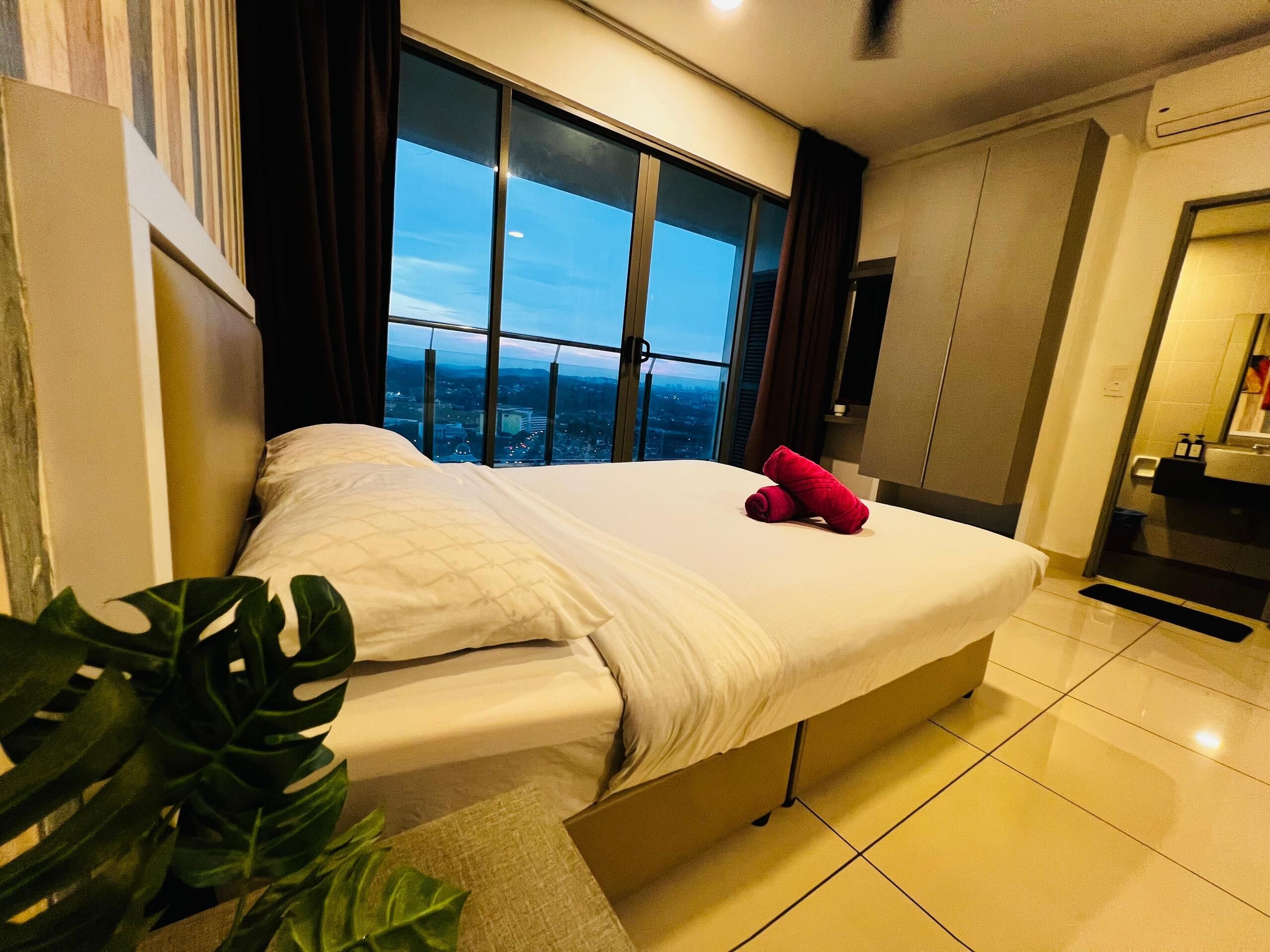 A hotel room with a large bed, a view of the city through the window, a plant, and a bathroom with a sink and mirror.