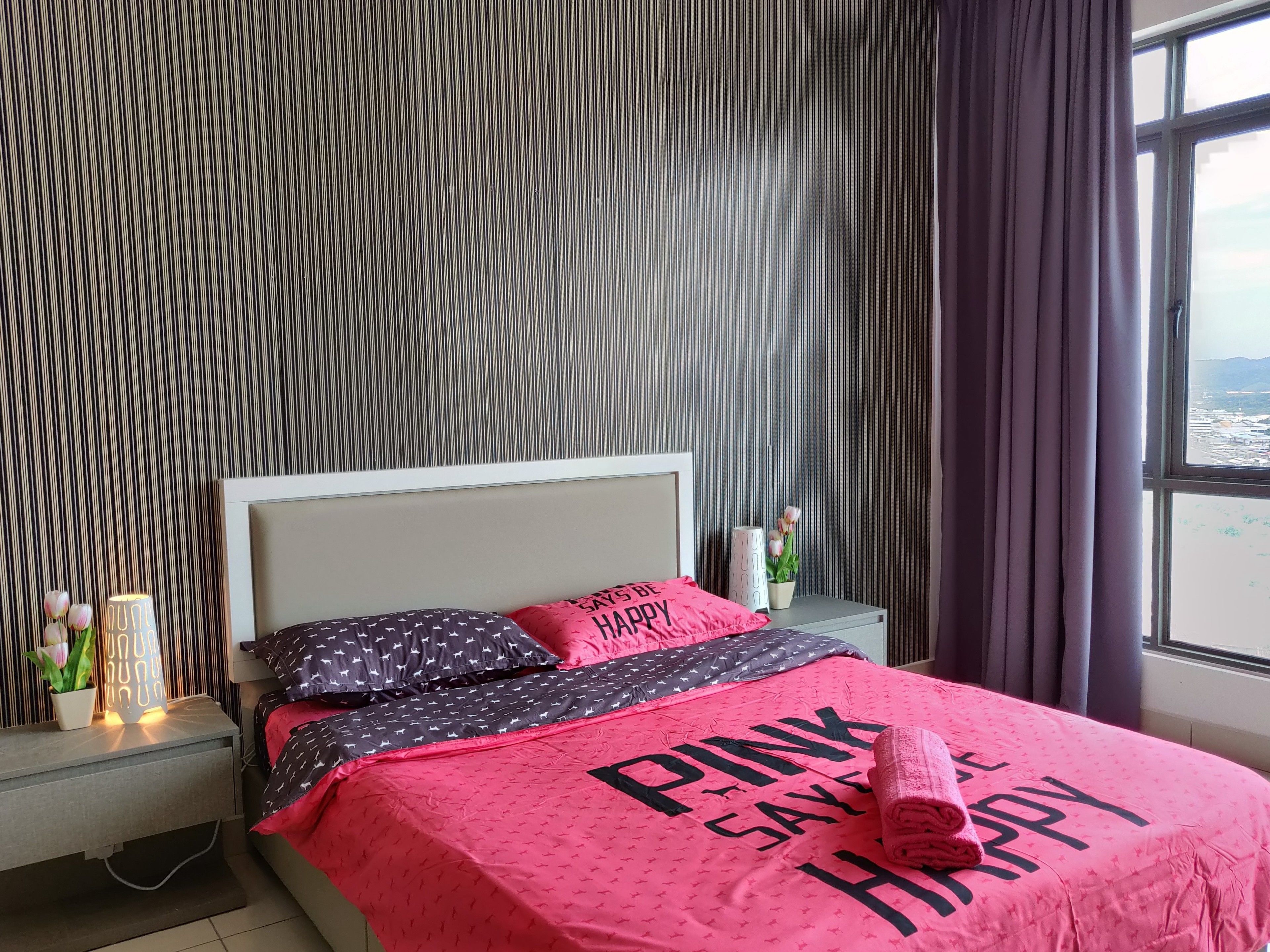 Una cama bien tendida con una colcha y almohadas rosas, una mesita de noche con una lámpara y una ventana con cortinas.