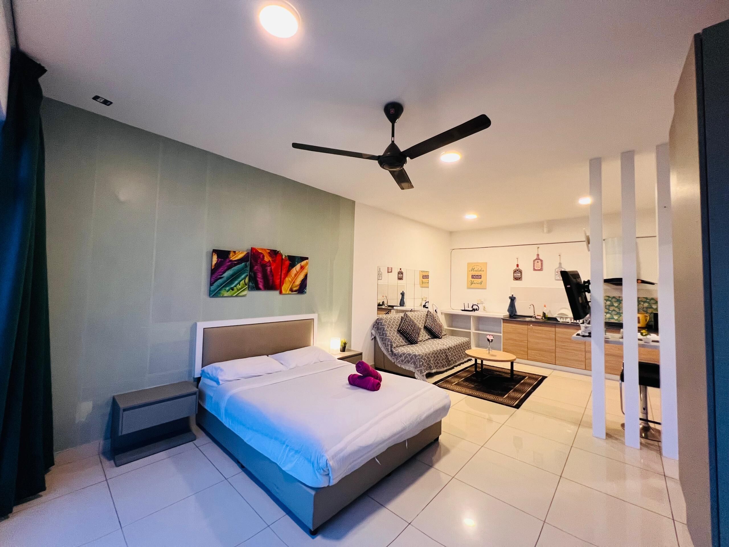 A modern hotel room with a bed, a ceiling fan, a small table, a chair, and a kitchenette in the background.