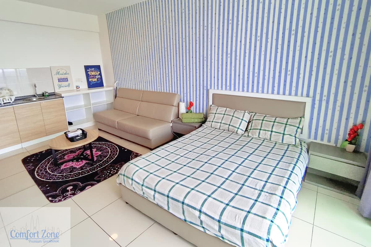 Un dormitorio con cama, sofá, mesita y mesita de noche. Hay una ventana con cortinas, una pared con diseño a rayas y una pared con un cartel.