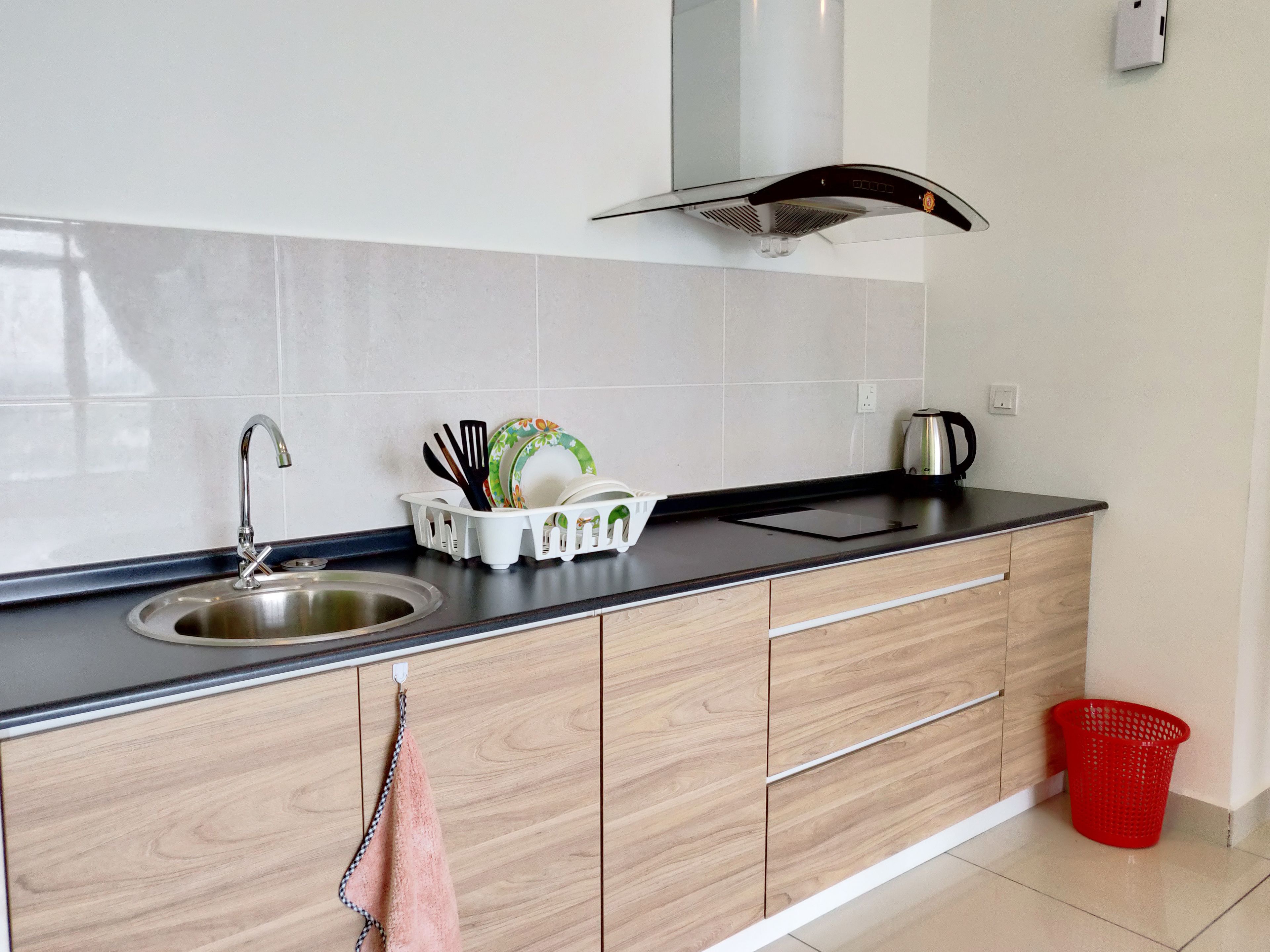 A modern kitchen with a stainless steel sink, a black countertop, wooden cabinets, a dish rack with utensils, and a red trash bin.