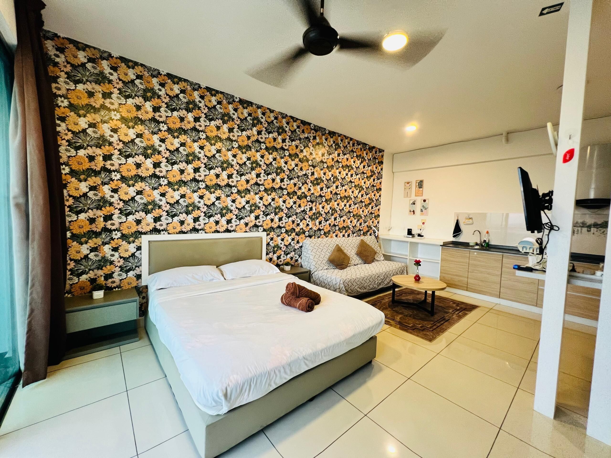 Quarto com cama, sofá, mesa pequena, ventoinha no teto e área de cozinha.