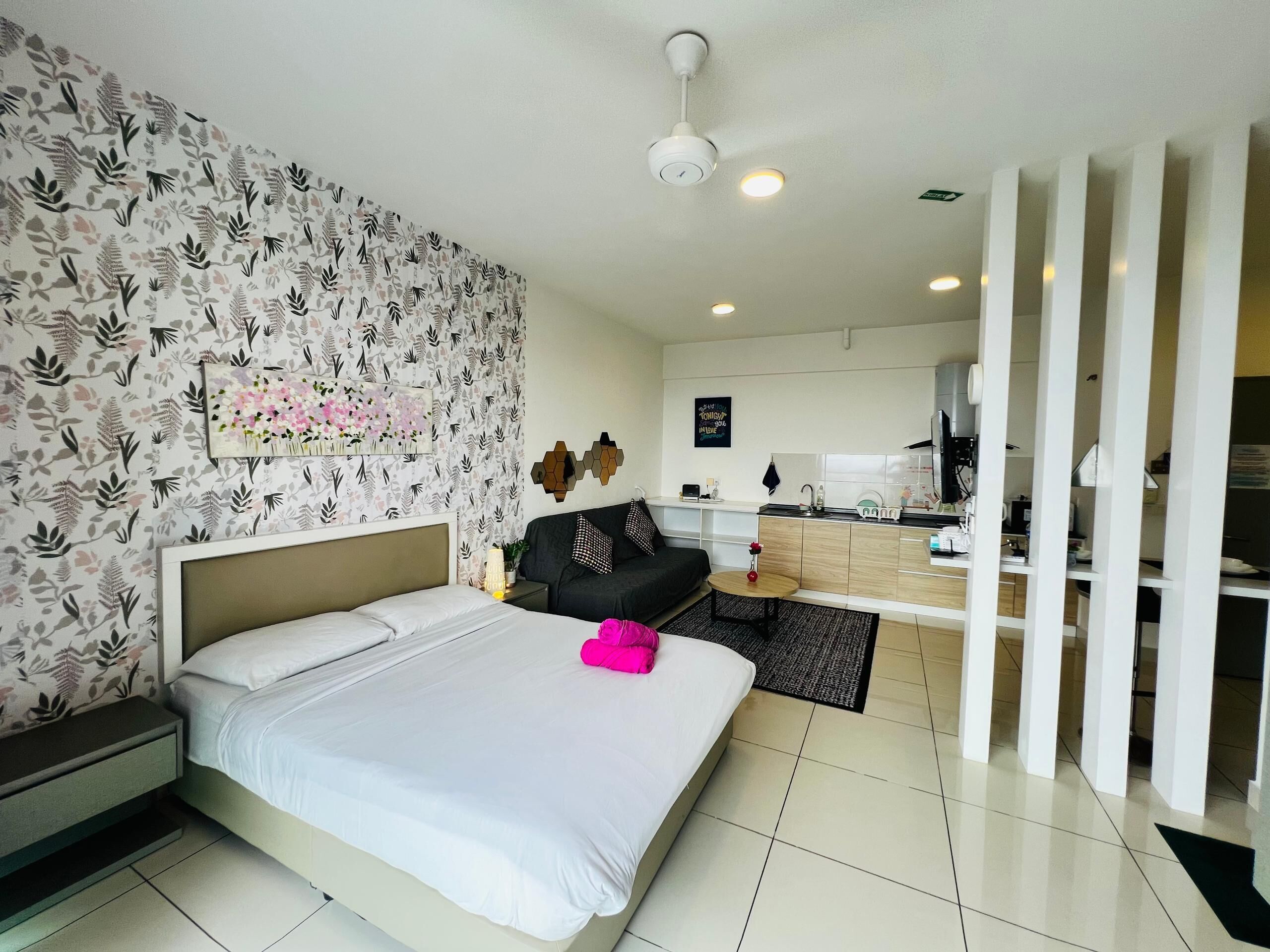 A modern hotel room with a bed, a small table with a vase, a sofa, a kitchen area, and a decorative wall.