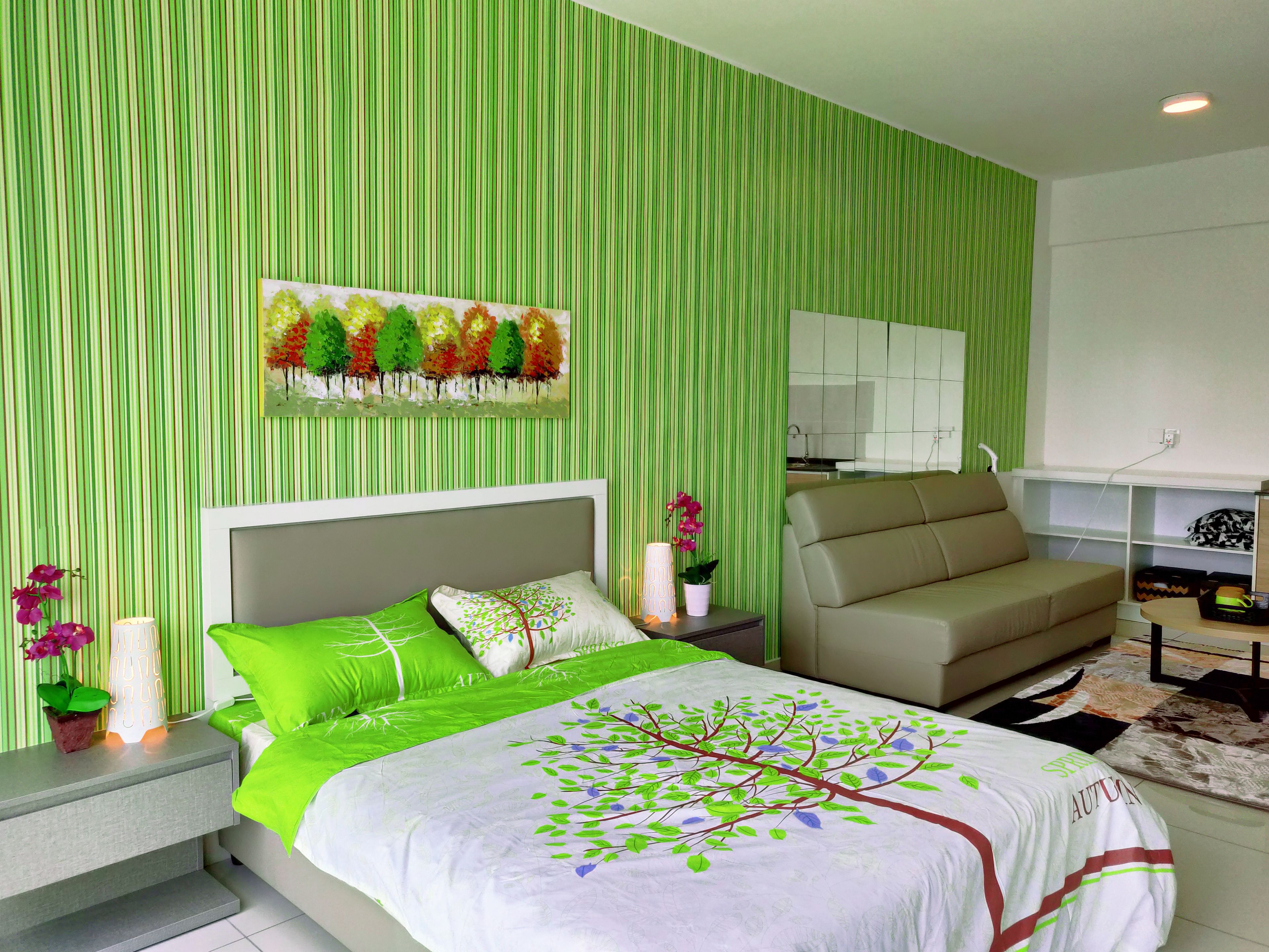Une chambre à coucher moderne avec un lit, un papier peint à rayures vertes, un tableau représentant des arbres, un canapé et une petite table avec une lampe et des fleurs.