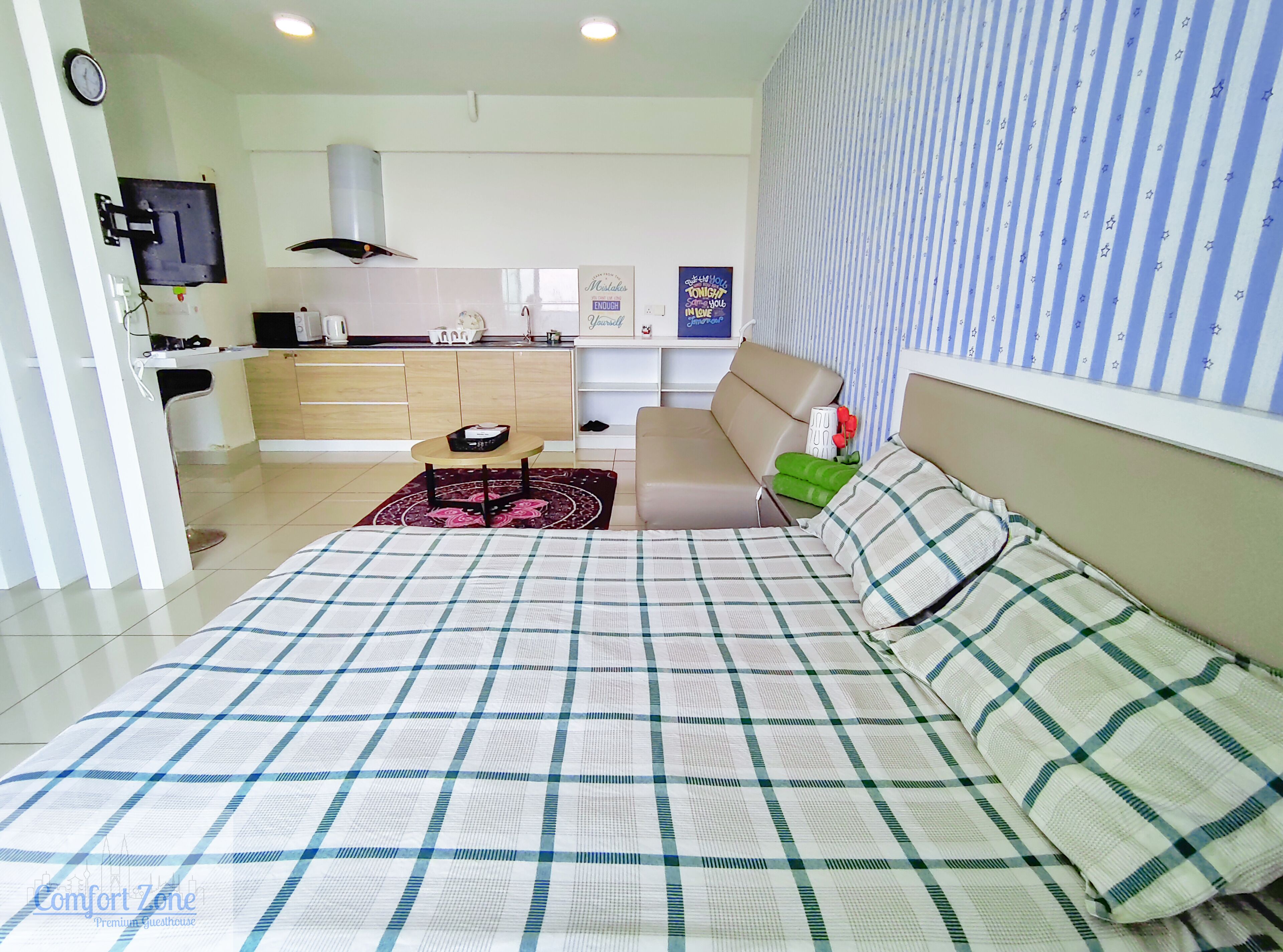 Un dormitorio con cama, sofá, una mesita y televisor. Se observa una cocina al fondo.