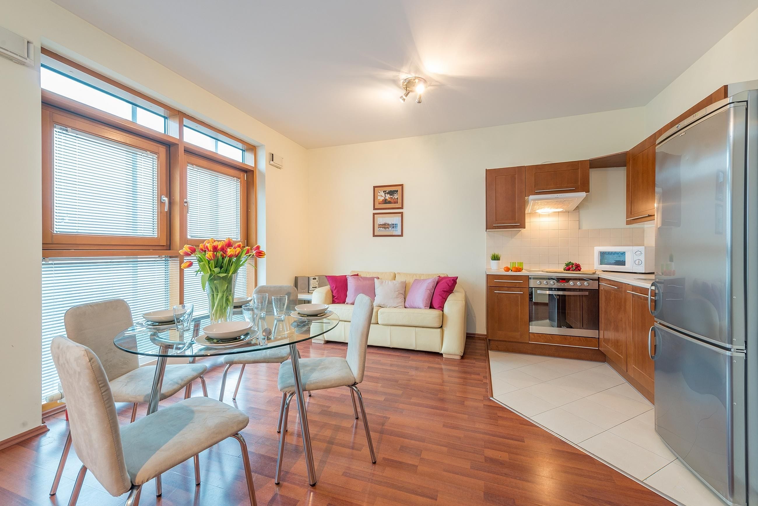 Una cocina moderna con armarios de madera, un refrigerador de acero inoxidable y una zona de comedor con mesa y sillas de vidrio.