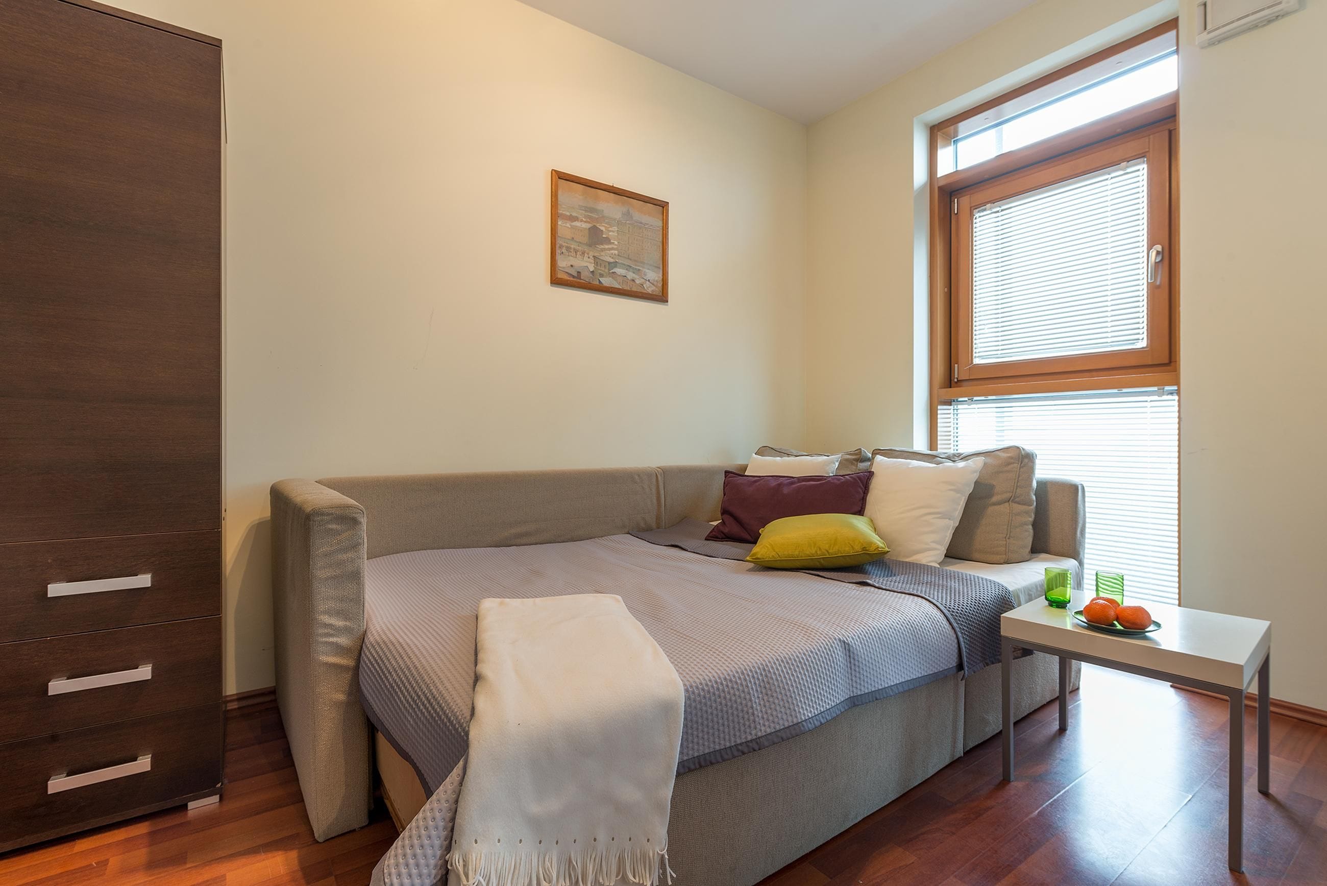 A bedroom with a bed, a nightstand, a window with blinds, a wardrobe, and a painting on the wall.
