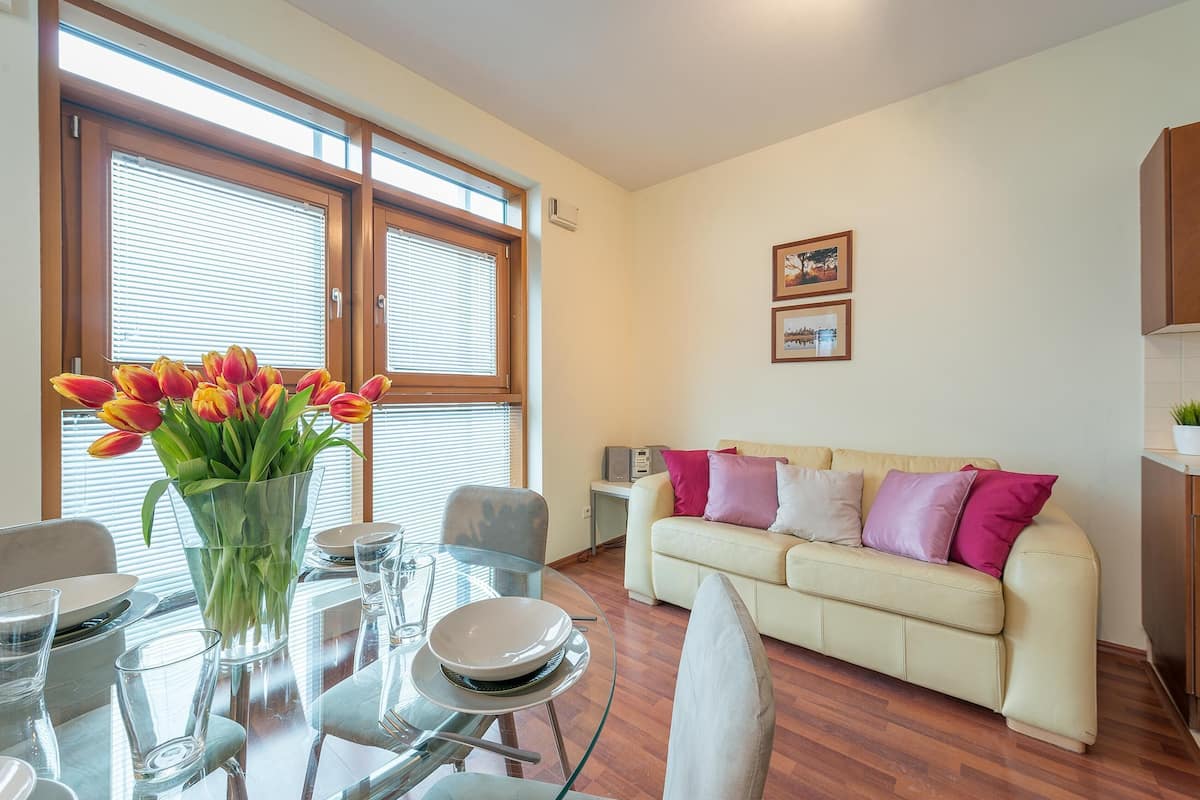 A dining area with a glass table, chairs, and a vase of tulips. There is a sofa with cushions and a small table with a speaker.