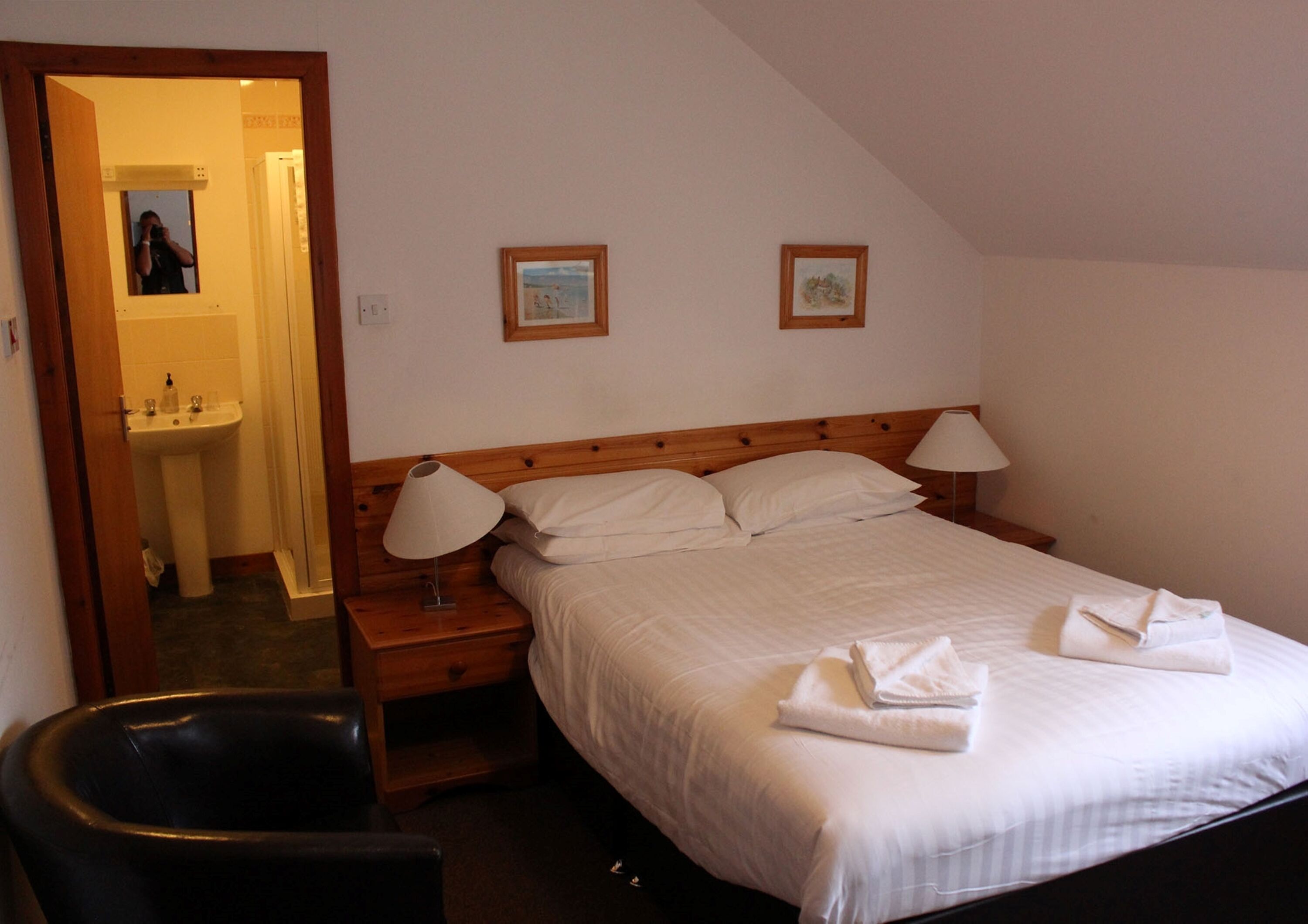 A bedroom with a bed, two bedside lamps, a wooden headboard, a nightstand, a chair, and a bathroom visible through an open door.