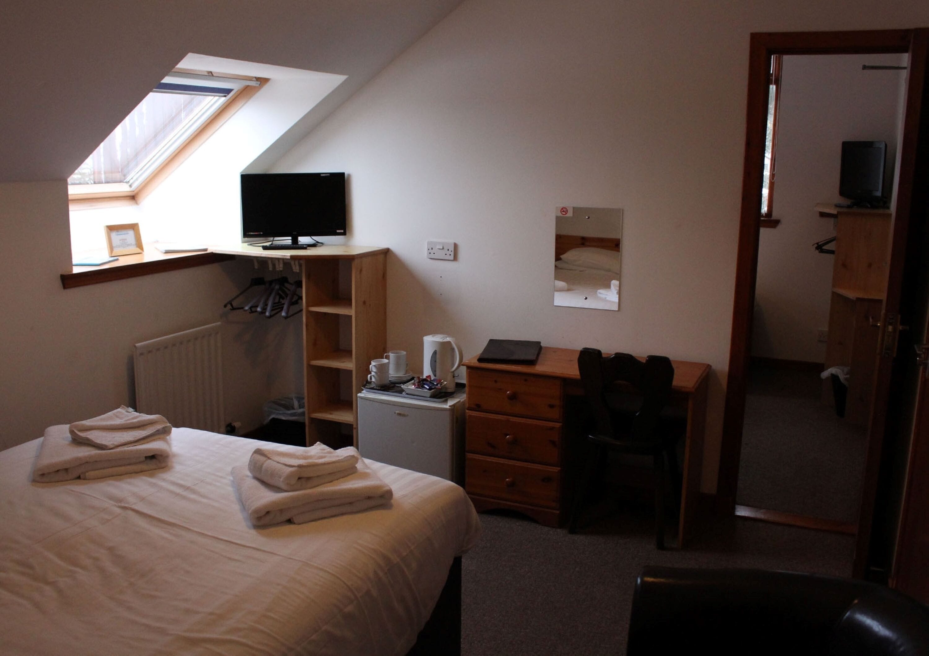 A bedroom with a bed, a desk, a chair, a radiator, a small refrigerator, and a television.