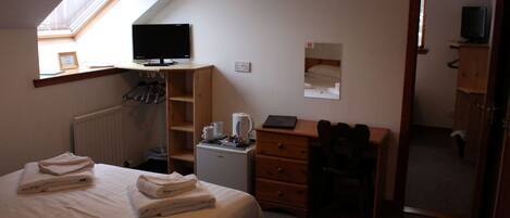 A bedroom with a bed, a desk, a chair, a radiator, a small refrigerator, and a television.