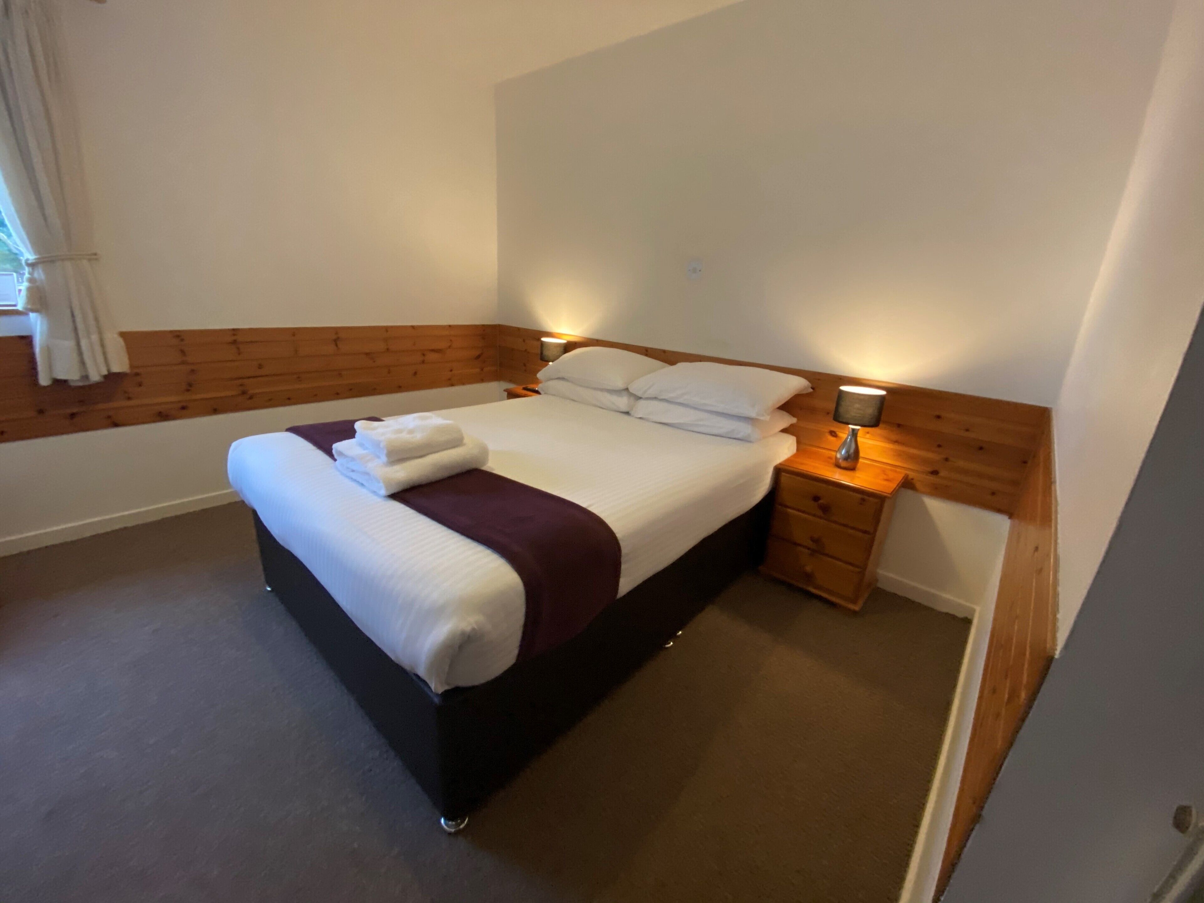 A neatly made bed with white linens and a purple throw, a bedside table with a lamp, and a wooden headboard.