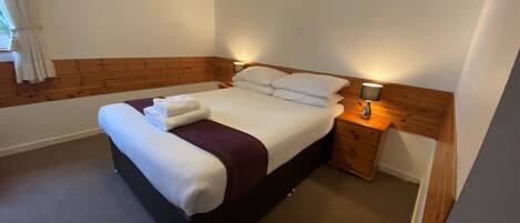 A neatly made bed with white linens and a purple throw, a bedside table with a lamp, and a wooden headboard.