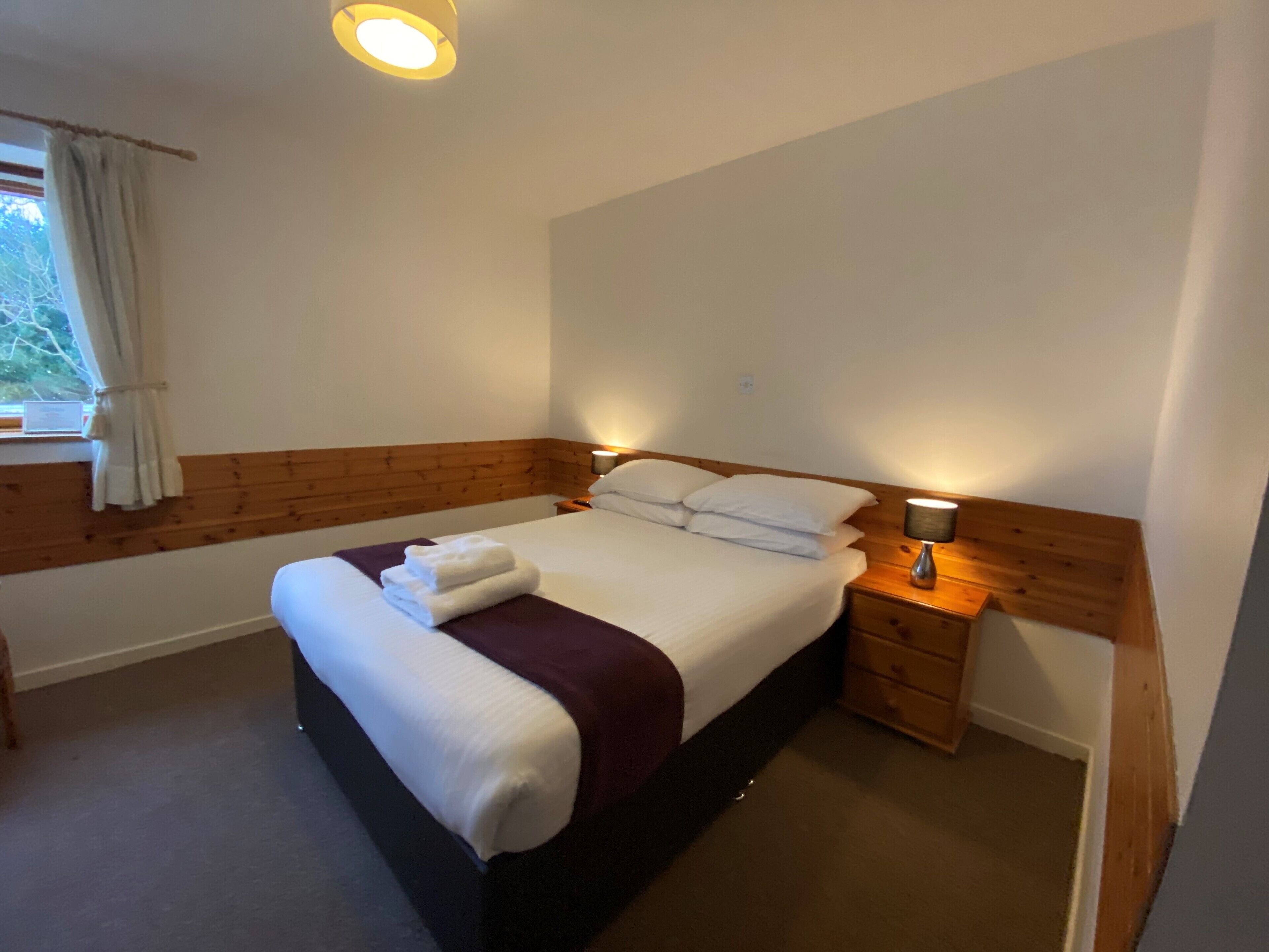 A neatly made bed with white linens and a brown headboard, a bedside lamp, and a window with curtains.