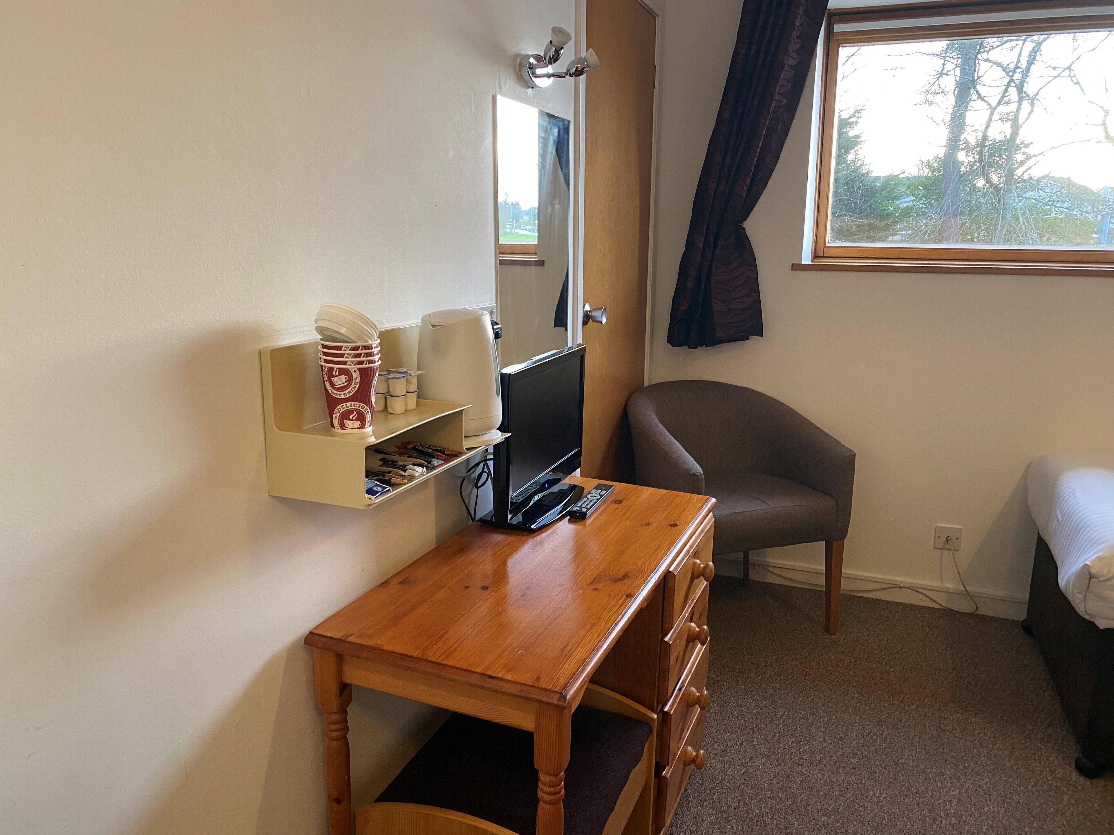 Une chambre d’hôtel avec un bureau en bois, une chaise, un lit, une fenêtre avec des rideaux et une étagère avec des tasses à café et une machine à café.