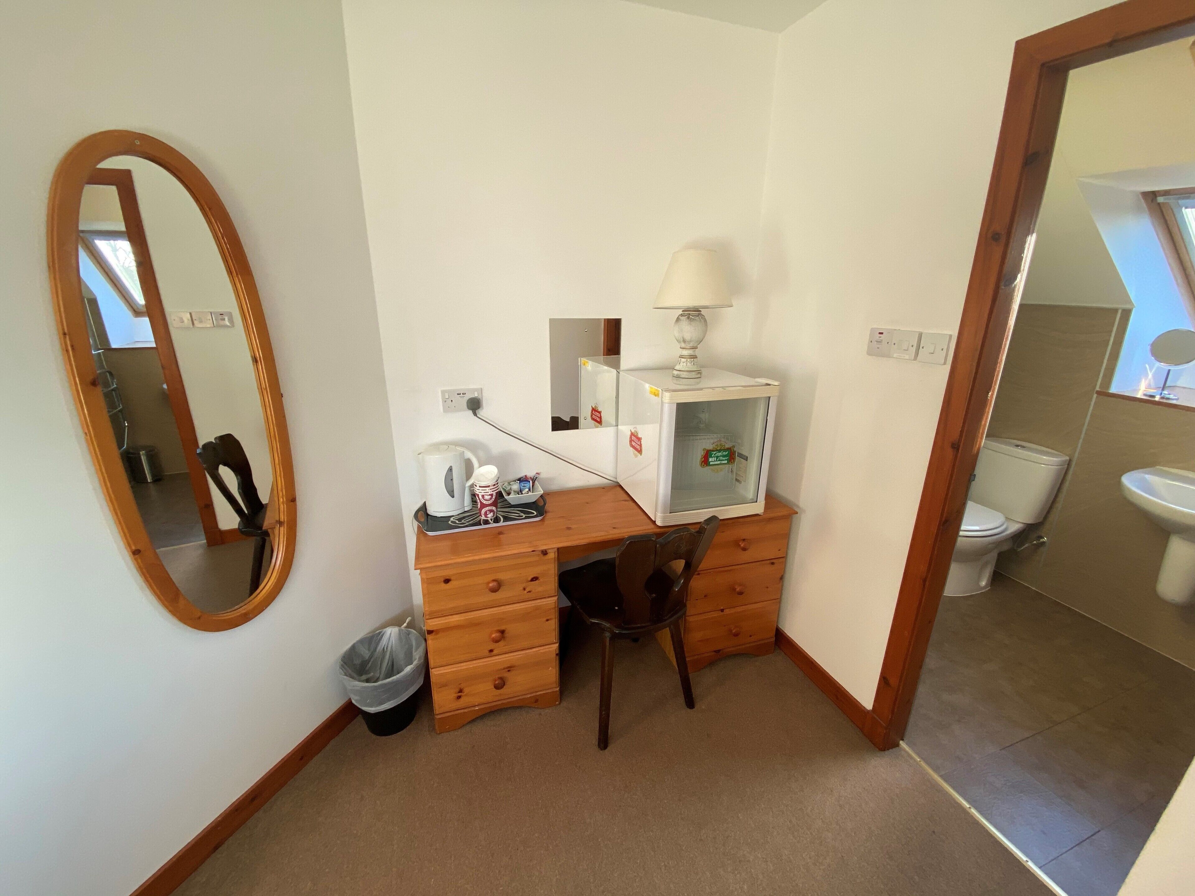 A compact bedroom with a wooden desk, a chair, a small trash bin, and a mirror.
