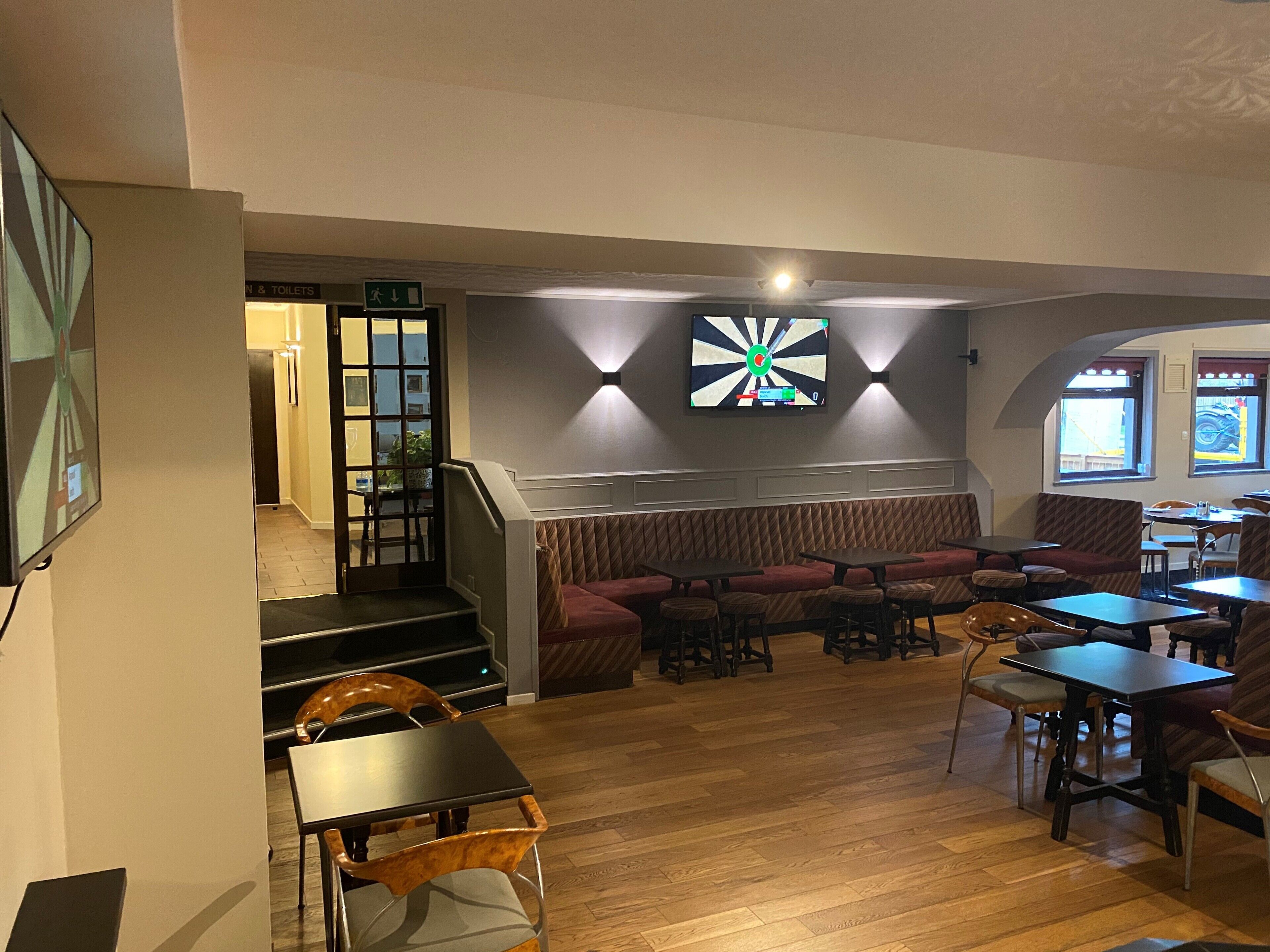 A restaurant with a wooden floor, seating area with tables and chairs, a staircase, and a dartboard on the wall.
