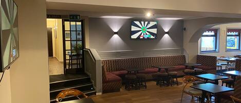 A restaurant with a wooden floor, seating area with tables and chairs, a staircase, and a dartboard on the wall.